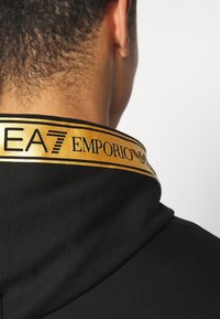 Svart plagg med en guldaccentuerad krage som har "EA7 EMPORIO"-märket, slät textur och minimalistisk design.