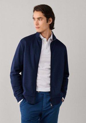 Junger Mann trägt eine marineblaue Jacke mit Reißverschluss über einem weißen Poloshirt und blauen Hosen, steht mit den Händen in den Taschen und schaut zur Seite.