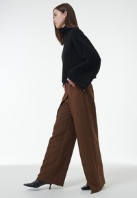 Un pull noir court avec un col montant et des manches larges, associé à un pantalon marron à taille haute et coupe ample, ainsi qu'à des escarpins noirs à bout pointu.