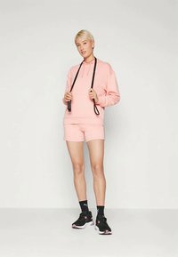 Ružová mikina s kapucňou a šnúrkou, spojená s kratšími nohavicami v rovnakej farbe. Čierne tenisky a ponožky po členky dotvárajú športový outfit. Hladký, ležérny materiál.