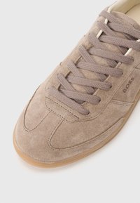 Sneaker in suede di un marrone chiaro con design curvo, punta rotonda, lacci beige e superficie testurizzata; presenta il nome del brand sul lato.