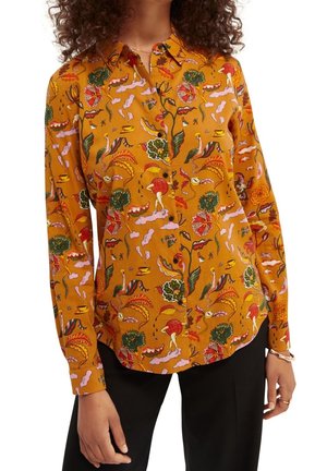 Frau trägt ein senfgelbes Hemd mit bunten, abstrakten Blumen- und Figurenmustern, kombiniert mit einer schwarzen Hose.