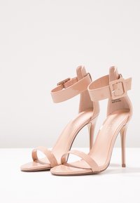 Nude patento-nahkasaappaat korkeilla koroilla, joissa on ohut nilkkanylky, avoin kärki ja tyylikäs stiletto-korko. Minimalistinen muotoilu sileällä pinnalla.