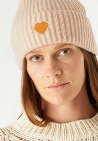 Lichtroze gebreide beanie met ribbelstructuur en een oranje hartvormige borduursel aan de voorkant, gedragen met een crèmekleurige gebreide trui.