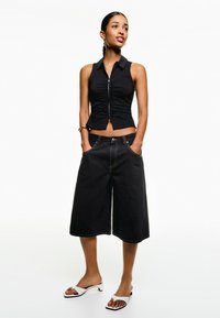 Black sleeveless top with front ruching and zip, paired with loose black culottes. White slip-on heels with a contrasting black sole.