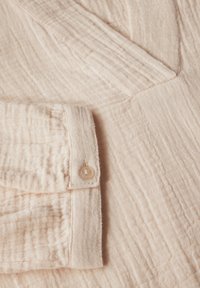 Textile pêche claire avec une texture froissée. Présente un poignet à boutons et un design en V. Le matériau semble doux et respirant.
