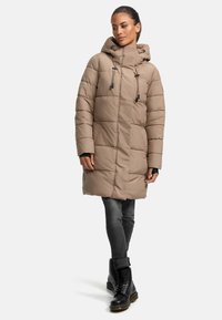 Abrigo acolchado beige con capucha, que presenta un diseño acolchado, detalles con cordones y bolsillos frontales, combinado con jeans oscuros y botas negras.