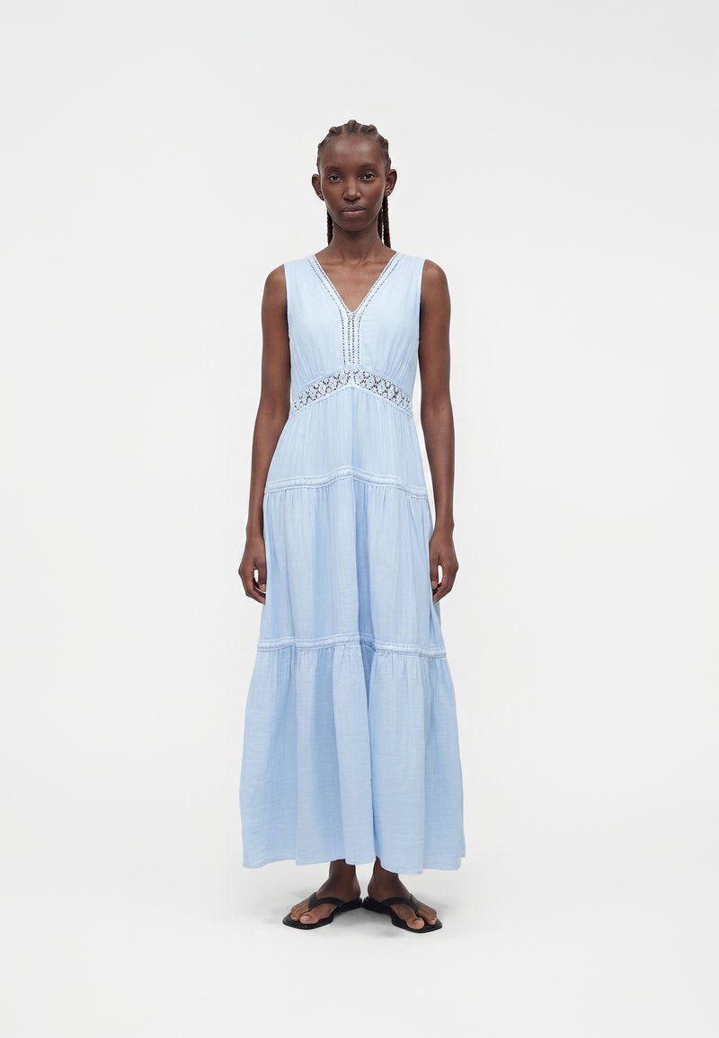 Vrouw met donker gevlochten haar die een mouwloze lichtblauwe maxi-jurk met kanten details draagt, staand op een effen witte achtergrond.