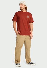 Homem vestido com uma t-shirt gráfica vermelha, calças bege, ténis pretos e brancos, e um gorro de malha branco, de pé com as mãos nos bolsos a olhar para a direita.