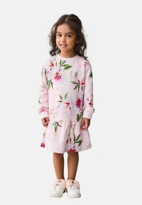 Rosa Blumenkleid mit langen Ärmeln, einem rüschierte Saum und goldenem Schriftzug. Kombiniert mit weißen Schuhen, die mit großen rosa Schleifen verziert sind.