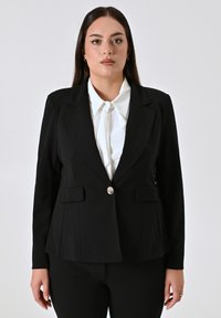 Zwarte op maat gemaakte blazer met een gestructureerd ontwerp, ingeknipte revers, sluiting met één zilveren knoop en twee voorzakken. Draagt over een witte blouse.
