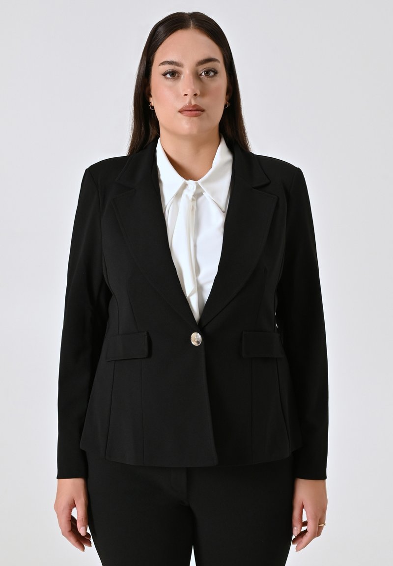 Zwarte op maat gemaakte blazer met een gestructureerd ontwerp, ingeknipte revers, sluiting met één zilveren knoop en twee voorzakken. Draagt over een witte blouse.