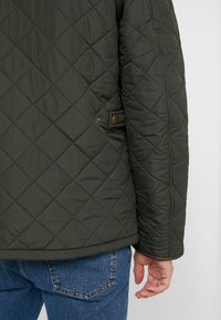 Quilted dark green jacket with a diamond pattern, featuring a side button detail. The fabric appears smooth and lightweight.