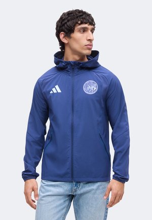 Man met een blauwe capuchonjas met een logo op de linkerborst en lichtblauwe spijkerbroek, kijkt naar rechts.