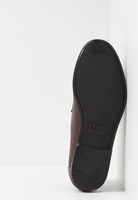 Chaussure en cuir marron avec une finition texturée et une semelle en caoutchouc noire. Elle présente un bout arrondi et un logo embossé sur la semelle.