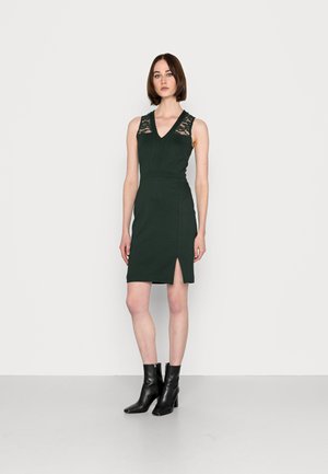 Vestido verde-escuro com detalhes em renda nos ombros, decote em V, silhueta ajustada e uma fenda lateral. Combinado com botas de tornozelo pretas.