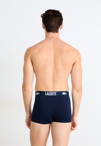 Jūriniai medvilniniai šortai su balta Lacoste logotipu ant juosmens, pasižymintys prigludusiu dydžiu ir lygia tekstūra, rodomi iš nugaros.