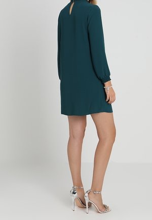 Kobieta w ciemnozielonej mini sukience z długim rękawem, nosząca srebrne sandały na wysokim obcasie i srebrną bransoletkę, stojąca na jednolitym tle.