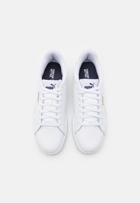 Baskets blanches en cuir synthétique avec un design à lacets, dotées d'une semelle intérieure bleu marine et d'un logo PUMA doré sur le côté. Texture lisse.