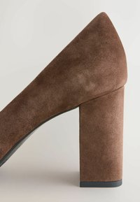Chaussure à talon haut en daim marron avec talon bloc et bout pointu, vue de côté sur un fond clair uni.