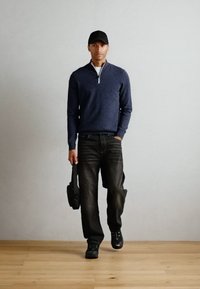 Navy zip-neck sweater, dark denim jeans, black cap, and shoes. Casual outfit with a fitted upper and relaxed lower silhouette. Carrying a shoulder bag.