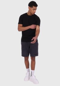 Sort bomulds t-shirt, grå fleece shorts, hvide høj-top sneakers og hvide sokker med sorte branddetaljer. Stående position med let armjustering.