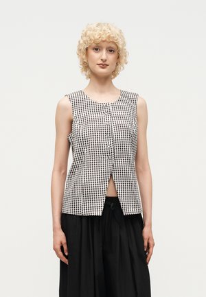 Personne aux cheveux blonds courts et bouclés portant un haut sans manches à carreaux noir et blanc boutonné et un pantalon noir ample.