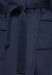 Tissu bleu marine avec une texture douce, présentant une ceinture nouée, des coutures visibles et une poche à rabat sur le côté gauche.