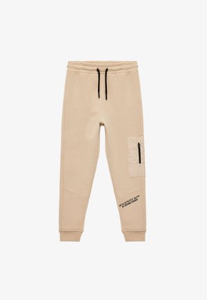 Pantalons de survêtement beiges avec une taille à cordon de serrage, des poignets élastiques, une poche latérale avec fermeture éclair et un détail en texte noir le long de la jambe. Texture douce.