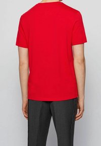 Person iført en ensfarvet rød kortærmet T-shirt og mørkegrå bukser, vist fra ryggen mod en lys grå baggrund.