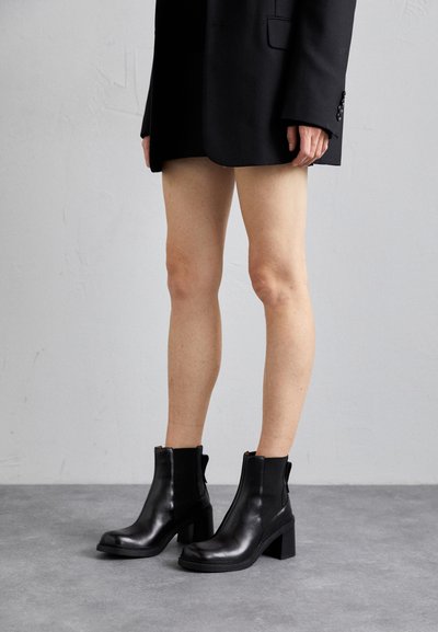 Botas negras hasta el tobillo hechas de cuero brillante, con punta cuadrada, tacón ancho, paneles elásticos laterales y una textura de superficie suave.