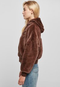 Haut à capuche en fausse fourrure marron avec une coupe décontractée, des poignets côtelés et une capuche doublée. Présente un logo brodé et une petite étiquette d'accent sur la manche.