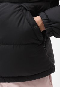 Czarna, pikowana kurtka o miękkiej teksturze, z elastycznymi mankietami oraz boczną kieszenią z zapięciem na zatrzask. Materiał wydaje się gładki i lekki.