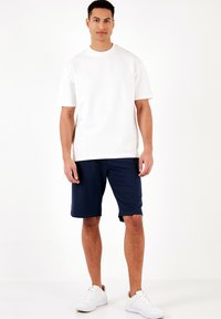 T-shirt bianco in cotone con maniche corte, abbinato a pantaloni corti blu navy. Modello che indossa sneakers bianche, in piedi su uno sfondo bianco uniforme.