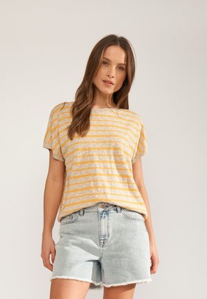 Femeie cu păr lung și castaniu, purtând un tricou cu mâneci scurte cu dungi galbene și bej și pantaloni scurți din denim albastru deschis, stând în picioare în fața unui fundal simplu.