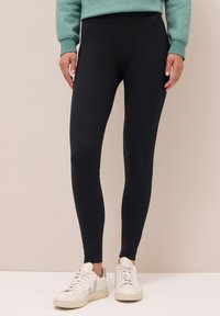 Leggings noirs en tissu extensible, dotés d'une taille haute et d'une texture lisse. Associés à des baskets blanches.