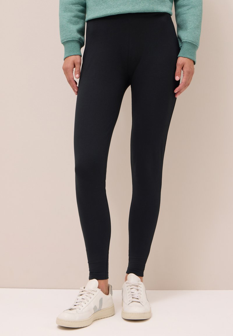 Leggings noirs en tissu extensible, dotés d'une taille haute et d'une texture lisse. Associés à des baskets blanches.