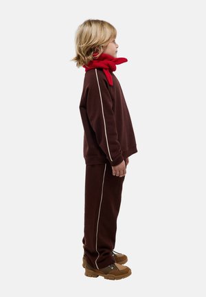 Enfant en survêtement marron avec des bandes blanches, debout de profil, portant une écharpe rouge et des bottes de randonnée marron clair sur un fond uni.