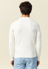 Maglione bianco a maglia con scollo rotondo e maniche lunghe, caratterizzato da un motivo testurizzato e polsini e orlo a coste. Indossato con jeans azzurri chiari.