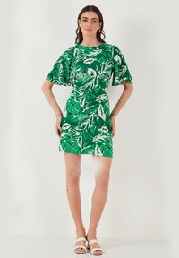 Vihreä mekko valkoisilla lehtikuvioilla. Lyhyet hihat ja tiukka muotoilu. Kankaan pinta näyttää olevan teksturoitu. Mallilla on valkoiset sandaalit.