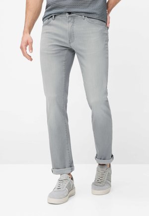 Mann trägt hellgraue hochgekrempelte Jeans, graue Sneakers und ein gemustertes Hemd, steht vor weißem Hintergrund.