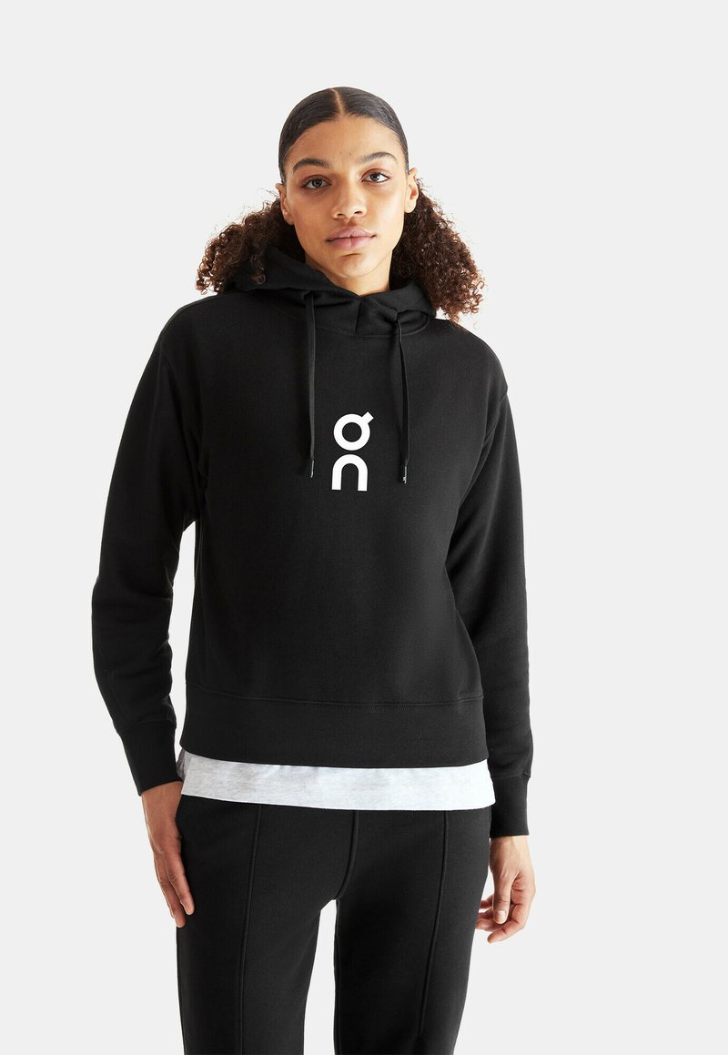 Sweat à capuche noir en mélange de coton, avec une poche avant, une capuche à cordon de serrage et un logo blanc. Associé à un pantalon sombre et un T-shirt gris.