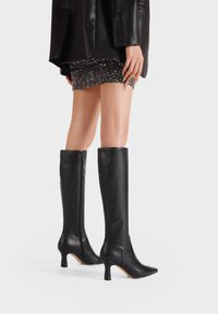 Stivali neri in pelle al ginocchio con punta affilata, tacco sottile e dettaglio con zip laterale, caratterizzati da una texture liscia e un design aderente.