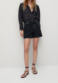 Femme portant une chemise noire à boutons, un short en denim noir taille haute avec une ceinture tressée, et des sandales plates à lanières noires, se tenant devant un fond uni.
