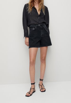 Woman wearing black button-up shirt, high-waisted black denim shorts with braided belt, and black strappy flat sandals, standing against plain background.