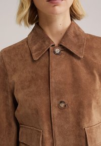 Bruine suède jas met een kraag, twee voorzakken en een knoopsluiting, met een gladde textuur en subtiele glans.