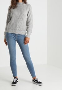 Grauer Strickpullover mit Chevron-Muster, kombiniert mit hellblauen Skinny-Jeans und schwarzen Turnschuhen mit weißen Akzenten.