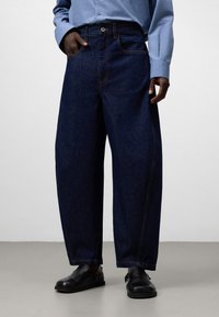 Mörkblå denimjeans med avslappnad passform, med framfickor, ljus sömnad och knappstängning. Bärs med svarta skor.