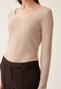 Haut beige à manches longues avec un col en V, en tissu tricoté texturé, coupe ajustée ; associé à un pantalon marron foncé avec un détail de boucle de ceinture en argent.