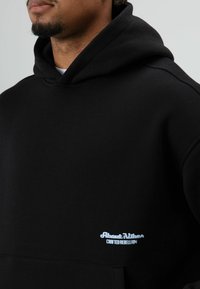 Zwarte hoodie van zachte stof, met een voorvak, een capuchon met trekkoord en een geborduurd logo in lichtblauw aan de linkerkant.
